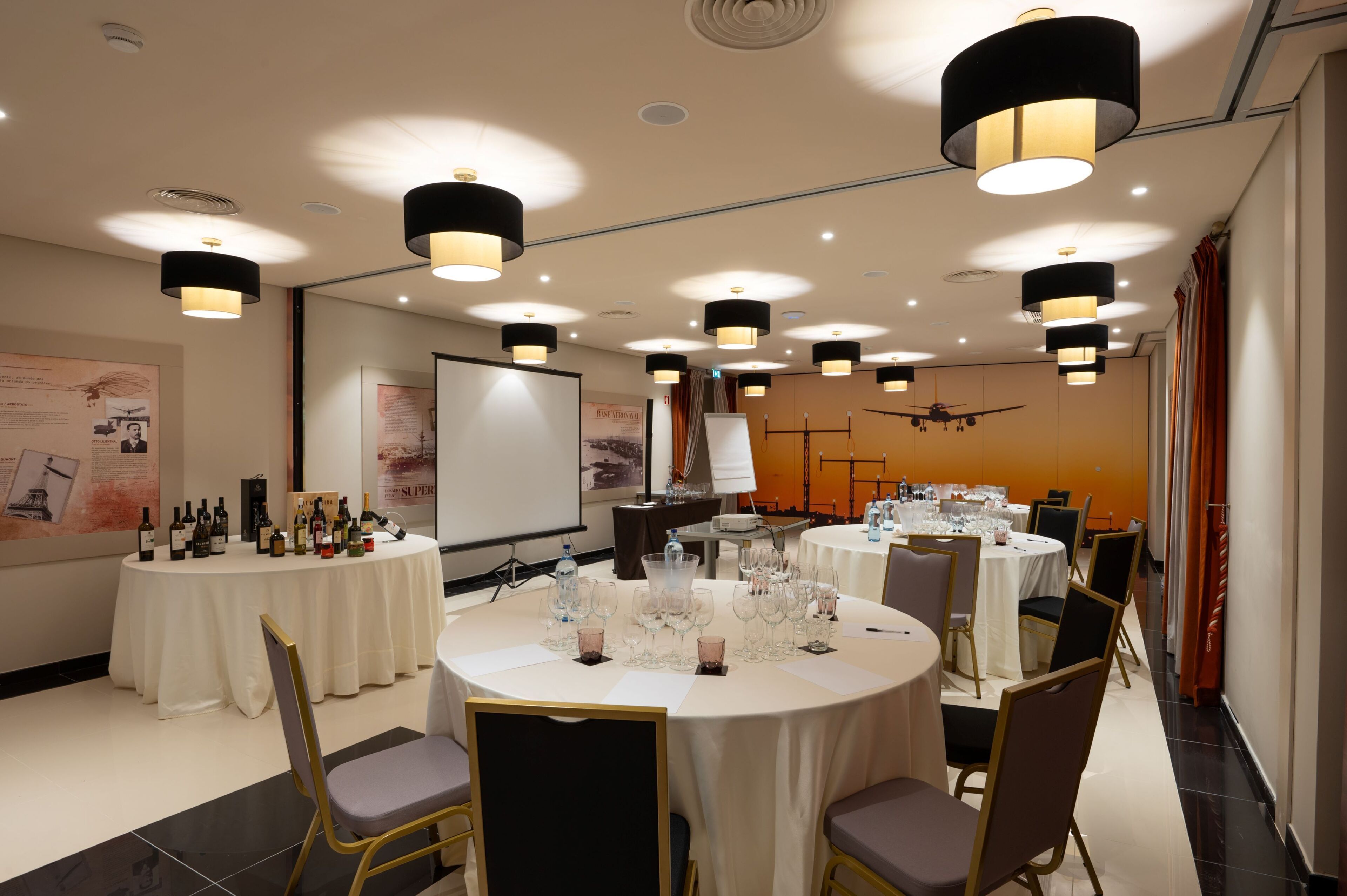 Un salón de conferencias con varias mesas redondas, cada una con sillas y vasos, y una pantalla de proyector al fondo.