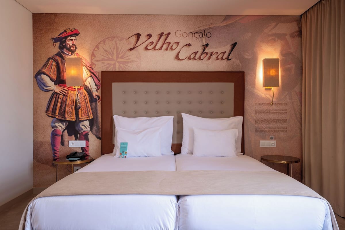 A hotel room with a large bed, two pillows, a nightstand, a wall-mounted lamp, and a mural of a historical figure.