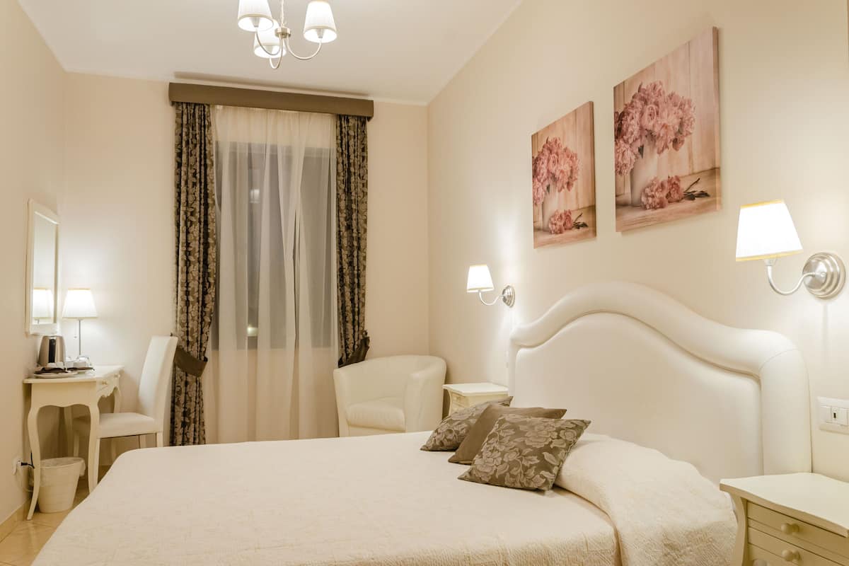 A hotel bedroom with a large bed, two bedside tables, a desk, a chair, and a window with curtains.