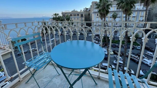 Un balcon avec une table et des chaises bleues, donnant sur un parking et des immeubles.