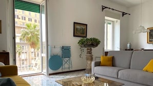 Un salon avec un canapé, une table basse en verre et un fauteuil. Une fenêtre offre une vue sur les bâtiments à l’extérieur.