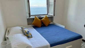 Un lit recouvert d’une couverture bleue, avec des oreillers blancs et un coussin jaune. Une fenêtre se trouve au-dessus du lit et offre une vue sur la mer.