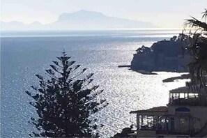 Une vue sur la côte, avec un ciel dégagé, un arbre au premier plan et un bâtiment doté d’un balcon donnant sur la mer.