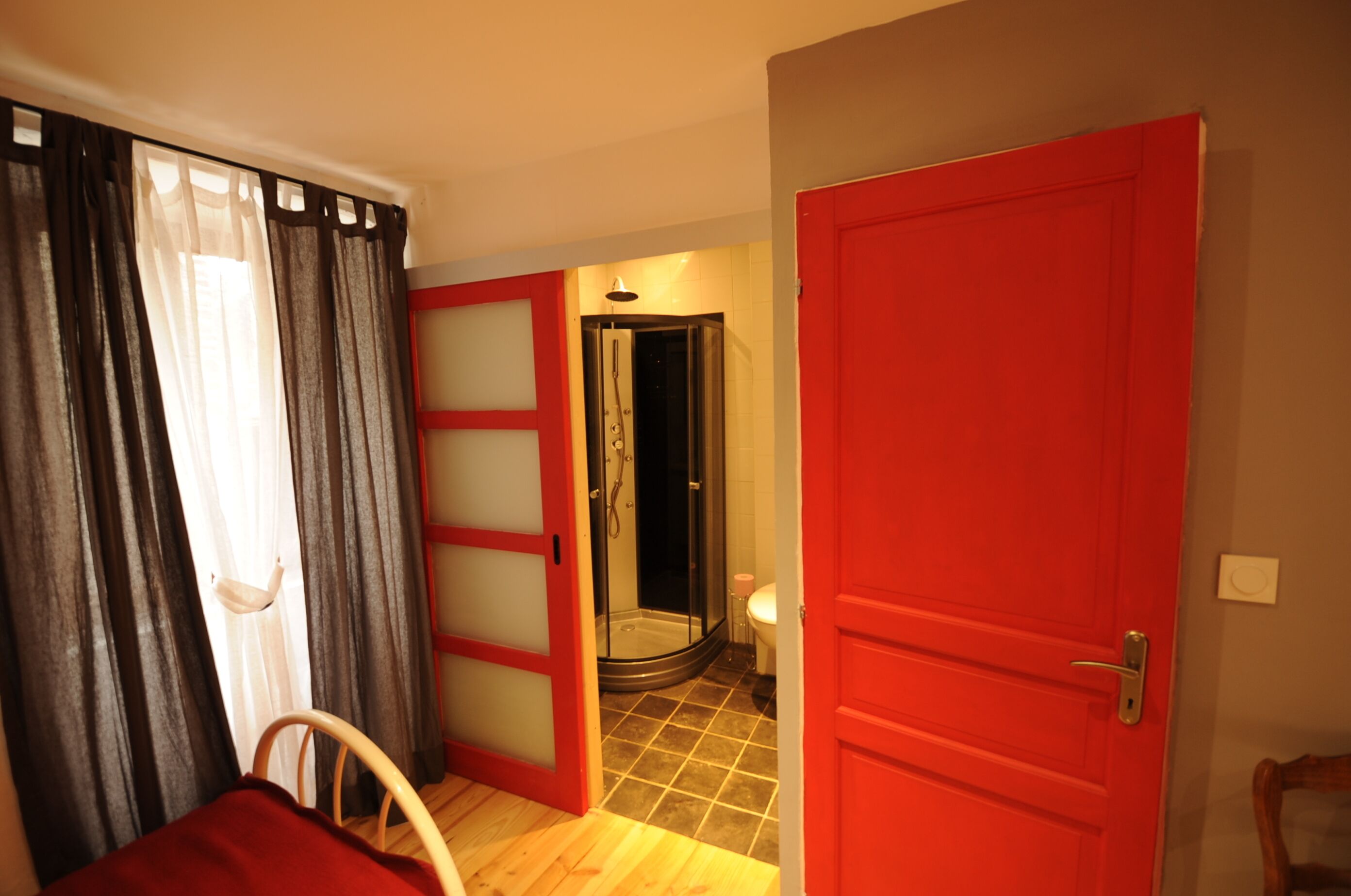 A hotel room with a red door, a shower area, and a view of the outside through a window with curtains.