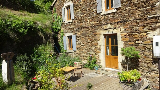 Un edificio de piedra con puerta y ventanas de madera, rodeado de vegetación y una terraza de madera.