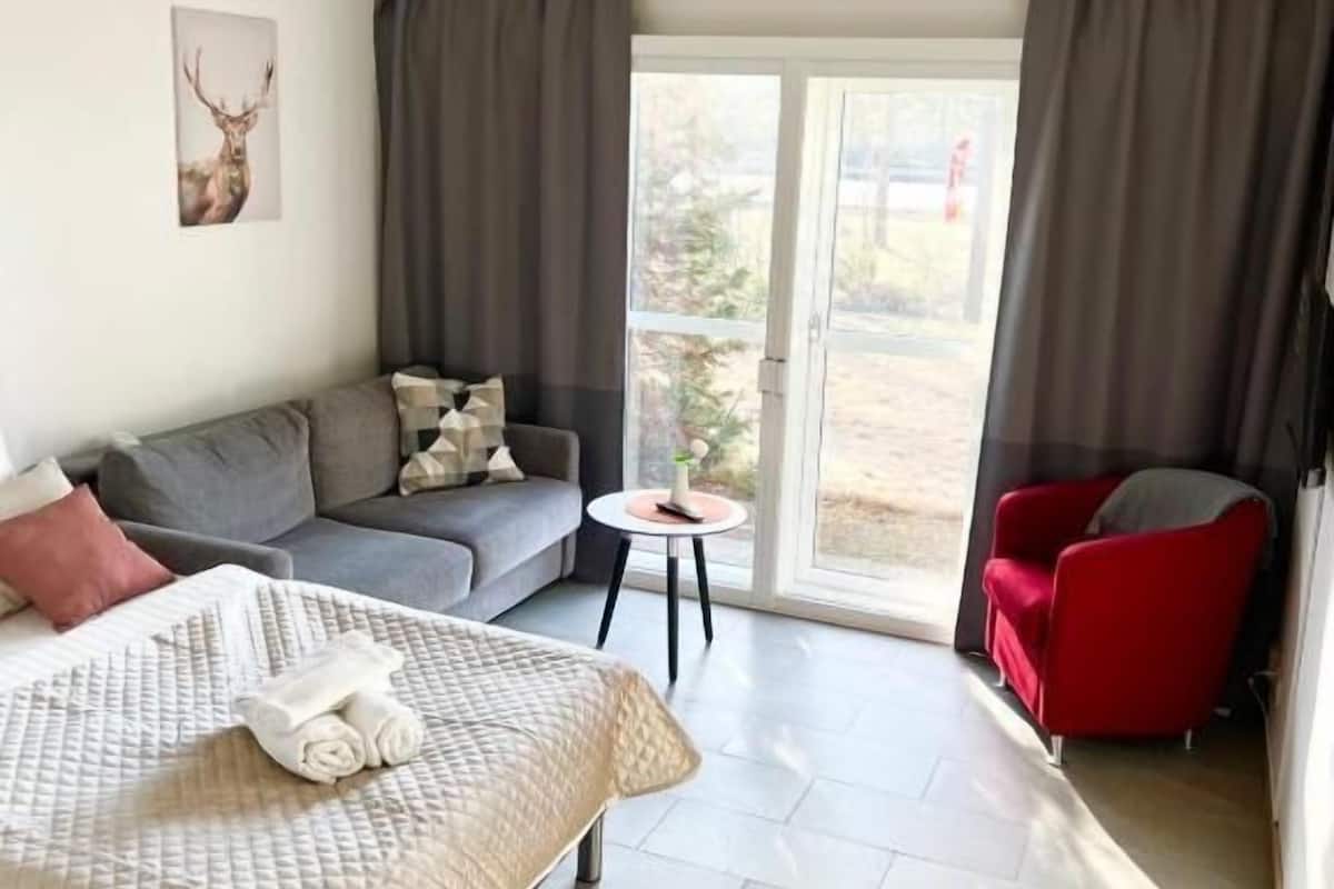 A modern hotel room with a bed, a grey sofa, a red armchair, and a small table.