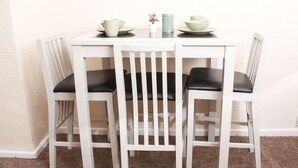 A white dining table with four chairs, each with a black cushion. There are two white cups and saucers on the table.