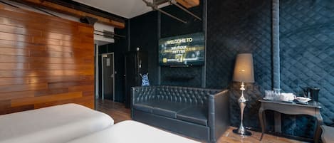 Ein modernes Hotelzimmer mit einer Wandpaneele aus Holz, einer schwarzen, gepolsterten Couch, einem Bett, einem Fernseher, der eine Willkommensbotschaft zeigt, und einem kleinen Tisch mit einer Lampe.