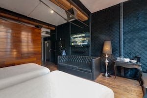 Una camera d'albergo moderna con una parete in legno decorata, un divano nero capitonné, un letto, una televisione che mostra un messaggio di benvenuto e un piccolo tavolo con una lampada.