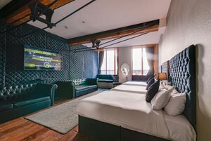 A modern hotel room with a large bed, a green sofa, a small table, and a wall-mounted TV displaying a welcome message.