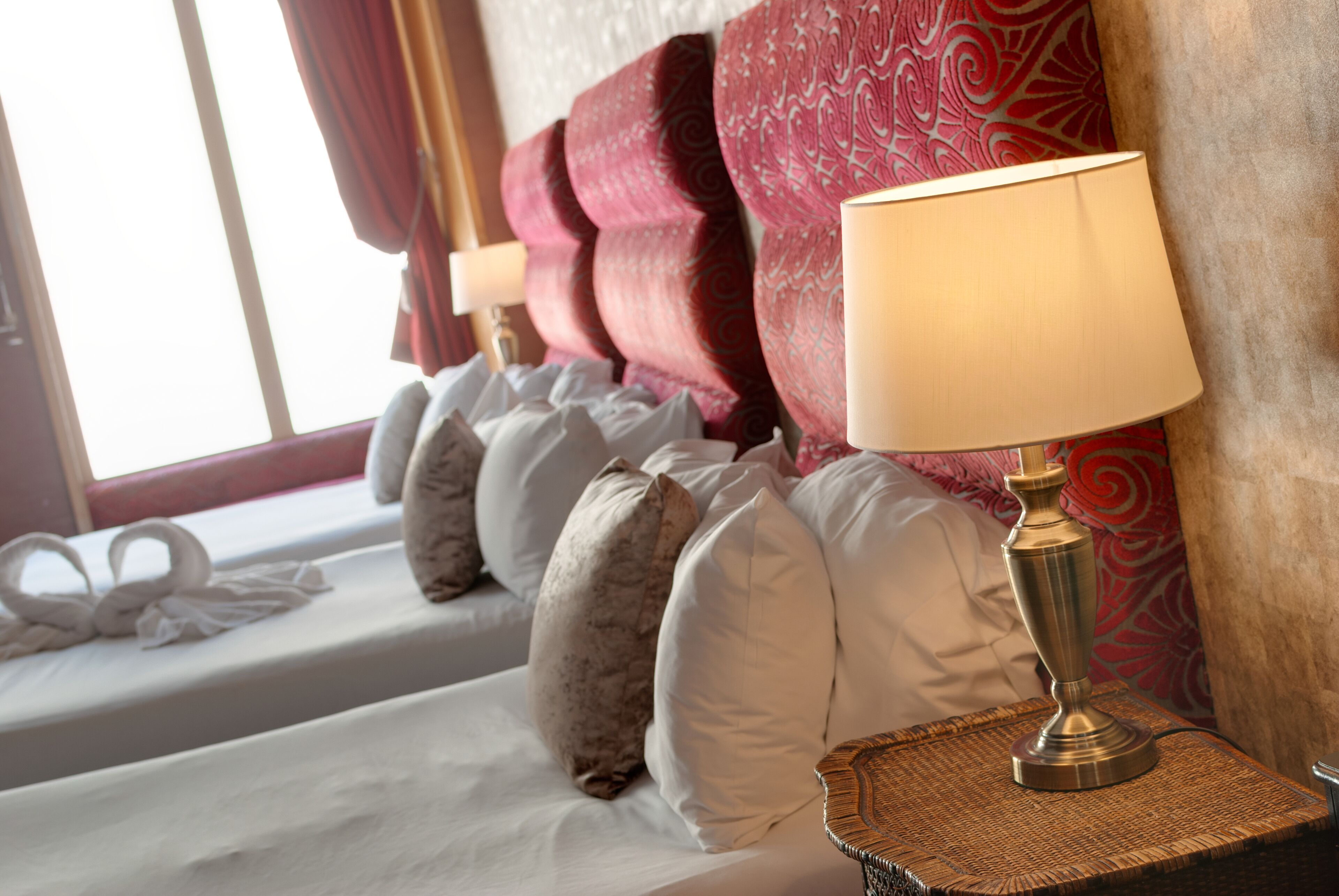 A neatly made bed with white and brown pillows, a decorative lamp on a woven tray, and a window with curtains.