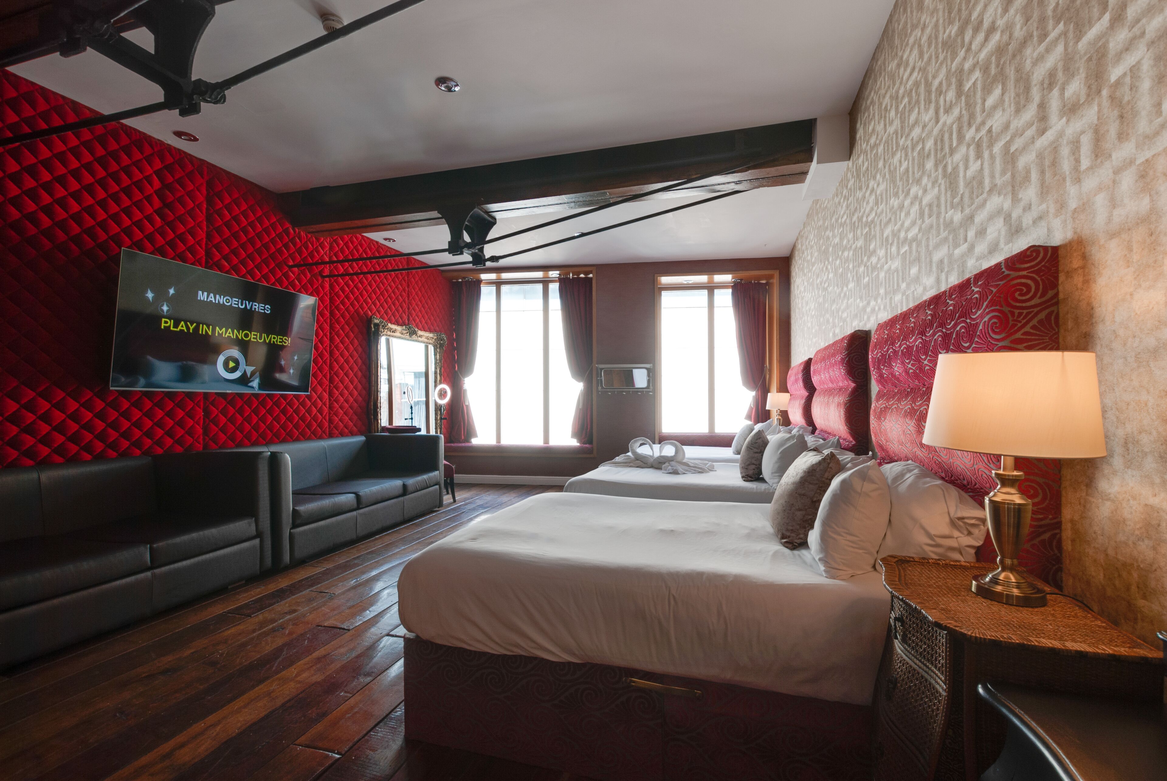 A hotel room with a red acoustic panel wall, a large flat-screen TV, a sofa, a bed with a headboard, and a lamp.