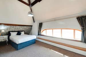 Un dormitorio con cama, mesita de noche, lámpara, ventana con cortinas y vistas al exterior.