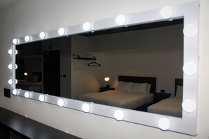 A hotel room with a large mirror with lights, a bed, a nightstand, and a hanging rack.