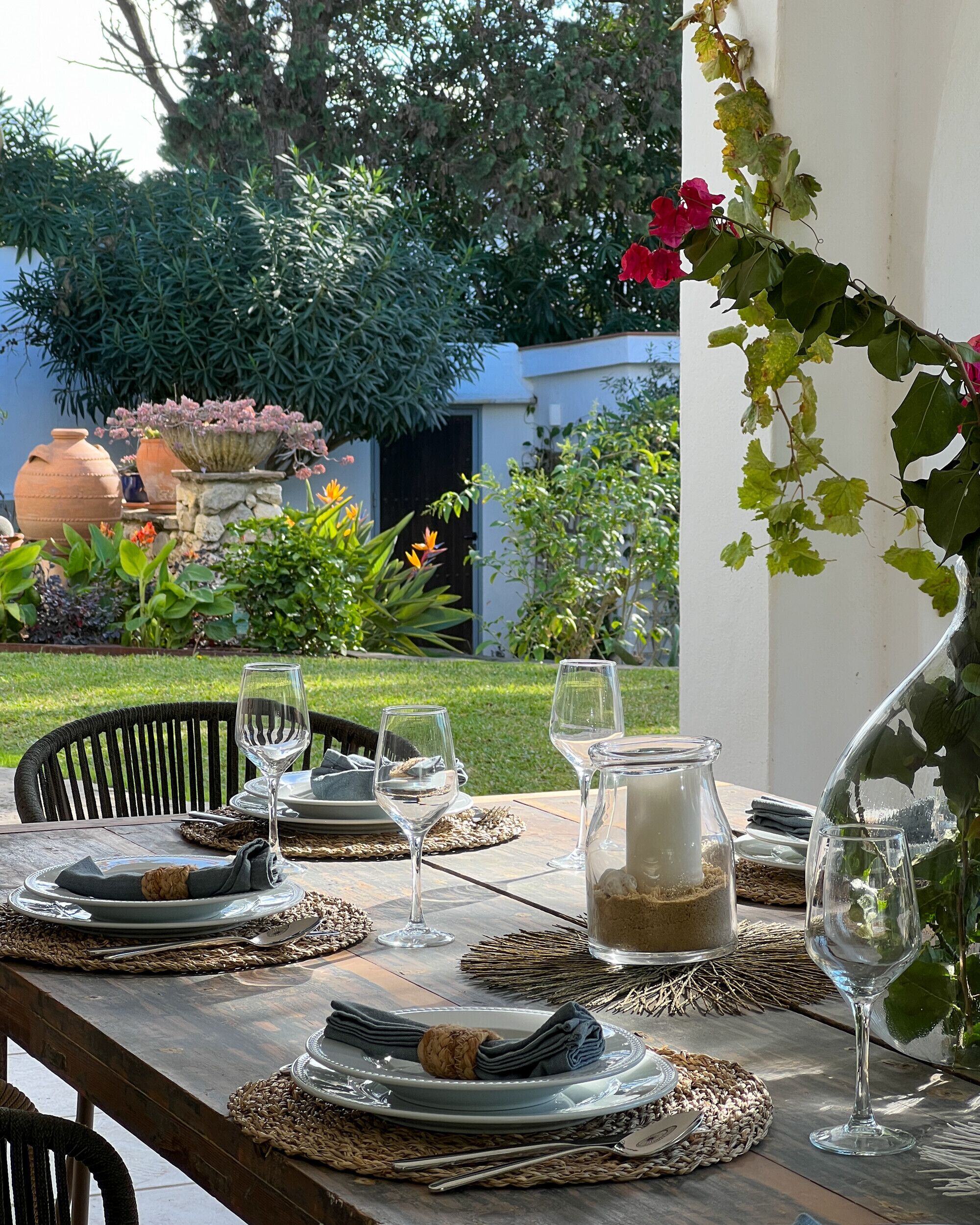 Un área de comedor al aire libre con una mesa de madera dispuesta para una comida, rodeada de follaje y un jardín.