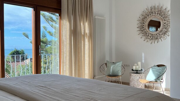 Ein Schlafzimmer mit einem großen Bett, einem Deckenventilator, Meerblick, einem runden Spiegel und einem Sessel mit Kissen.