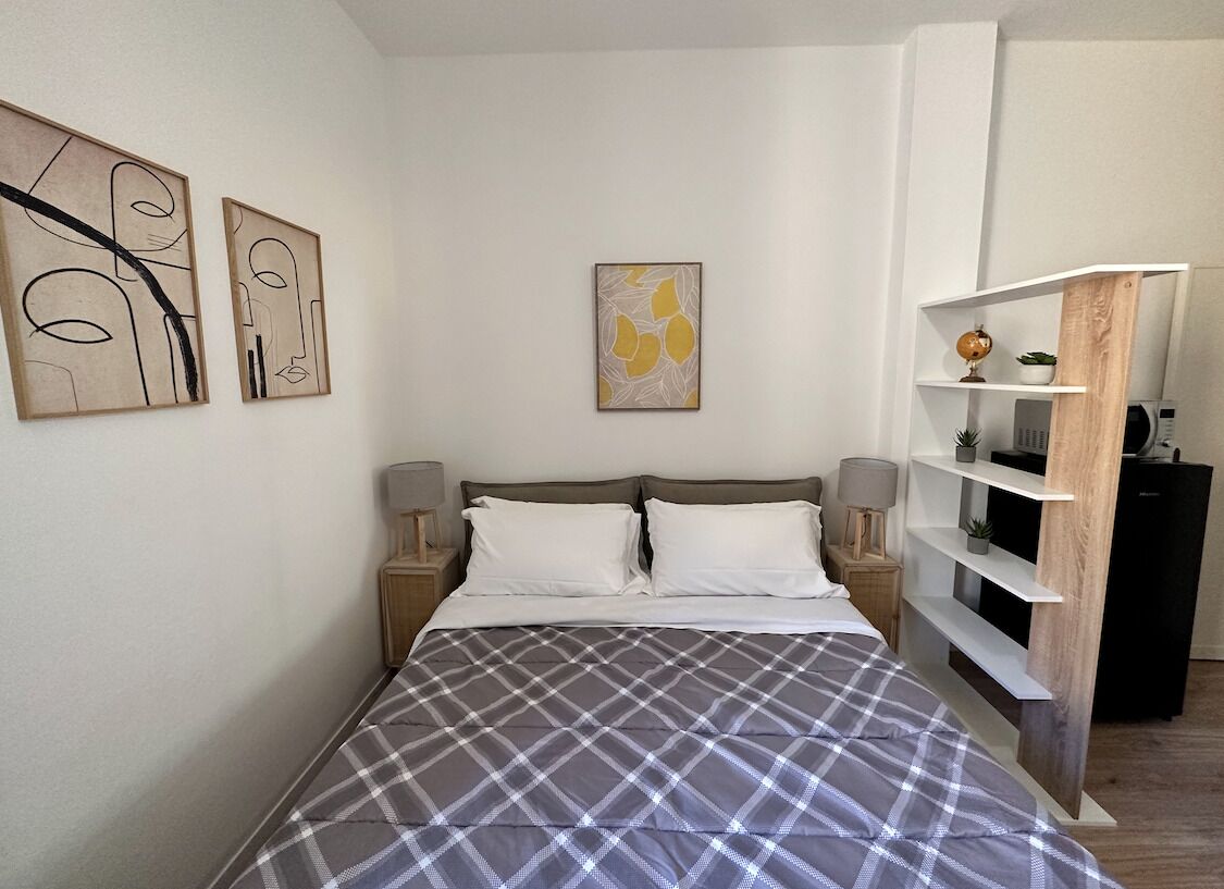 A neatly made bed with a plaid bedspread, a bedside table with a lamp, and a shelf with decorative items.