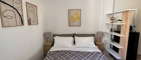Una cama bien hecha con cubrecama escocesa, una mesita de noche con lámpara y un estante con objetos decorativos.