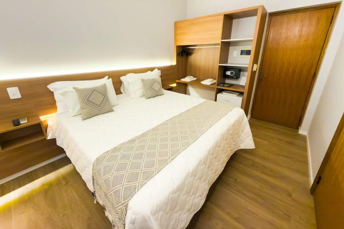 Cama bem arrumada com colcha estampada e almofadas, mesa de cabeceira com abajur, e guarda-roupa de madeira com espelho e prateleiras.