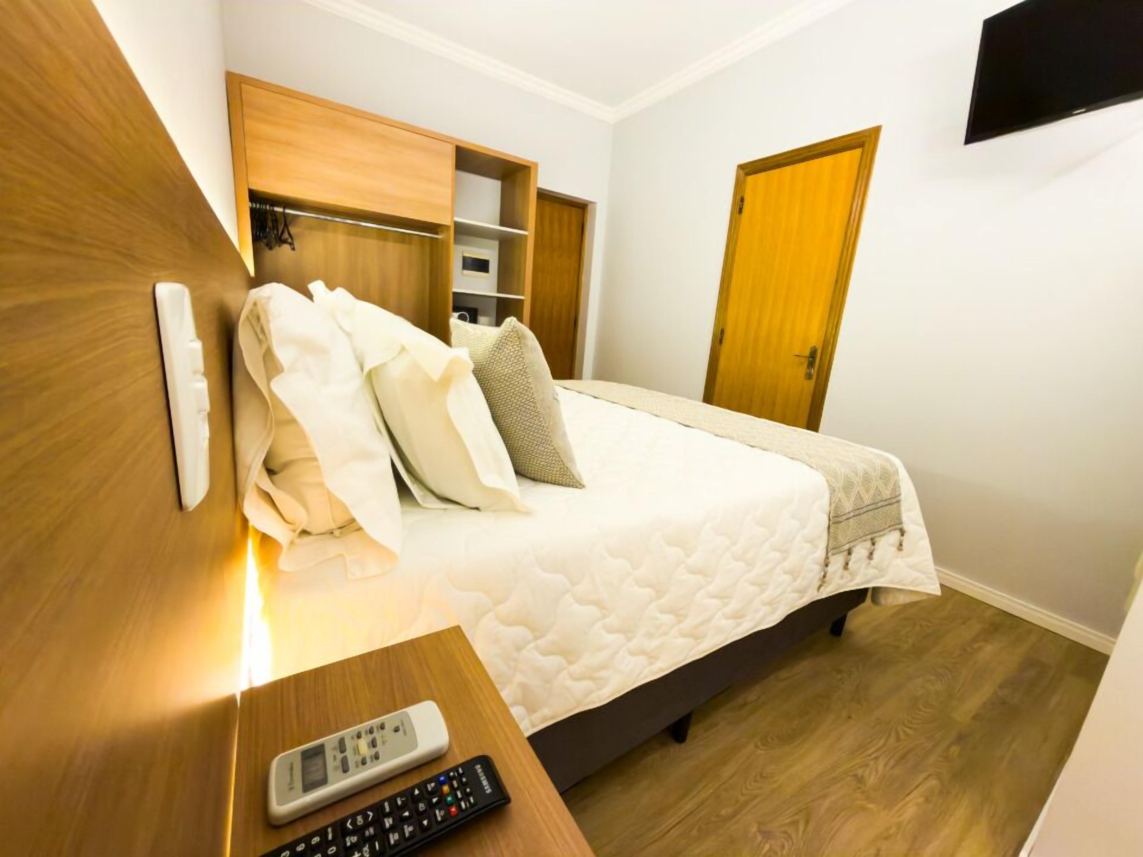 A hotel room with a bed, a bedside table with a remote control, a wooden wardrobe, and a television mounted on the wall.