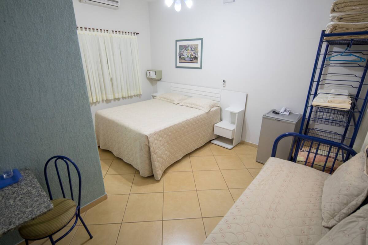 Um quarto com uma cama, uma cadeira, uma mesinha, um guarda-roupa e um quadro na parede.