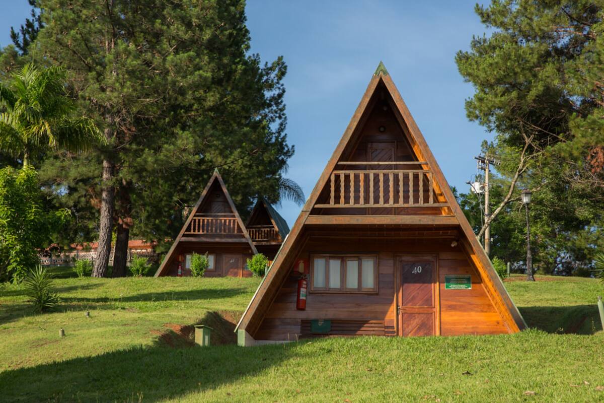 Duas cabanas de madeira com telhados triangulares, rodeadas por árvores e relva.