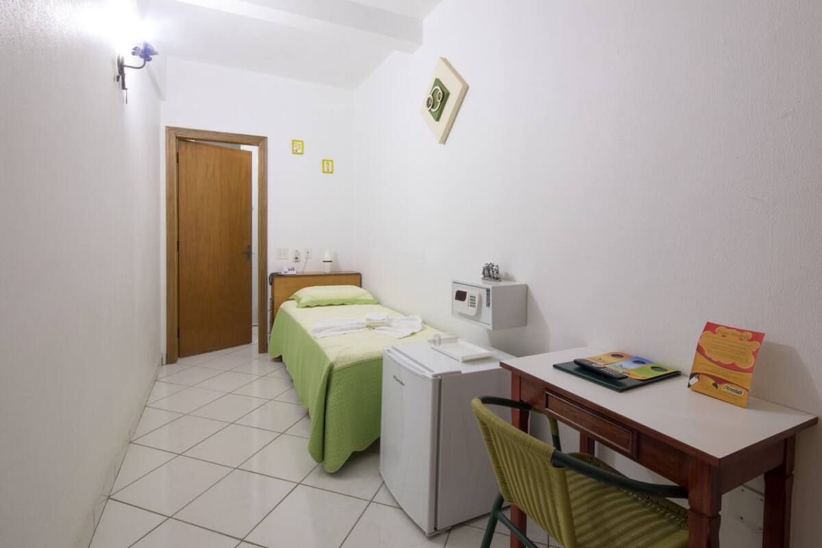Quarto pequeno e limpo, com cama, escrivaninha, cadeira, frigorífico e aquecedor fixado na parede.