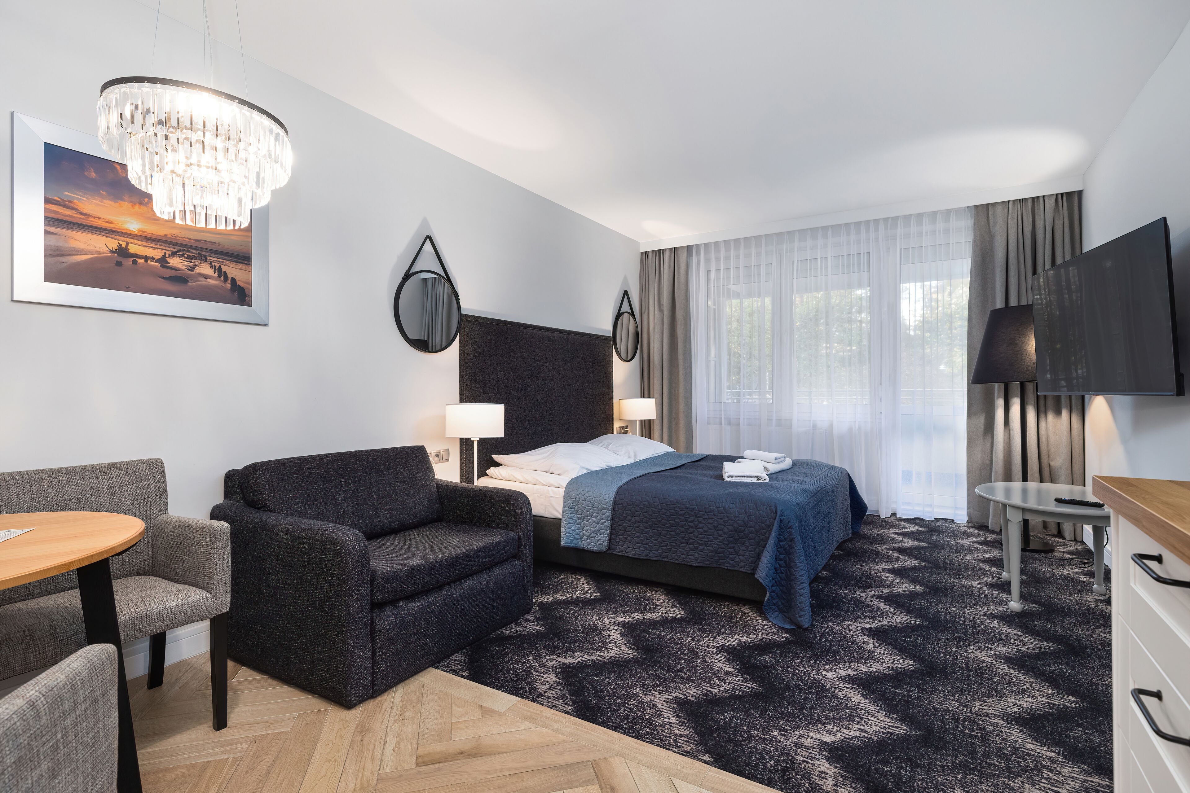 A modern hotel room with a bed, a sofa, a desk, a chandelier, and a television.