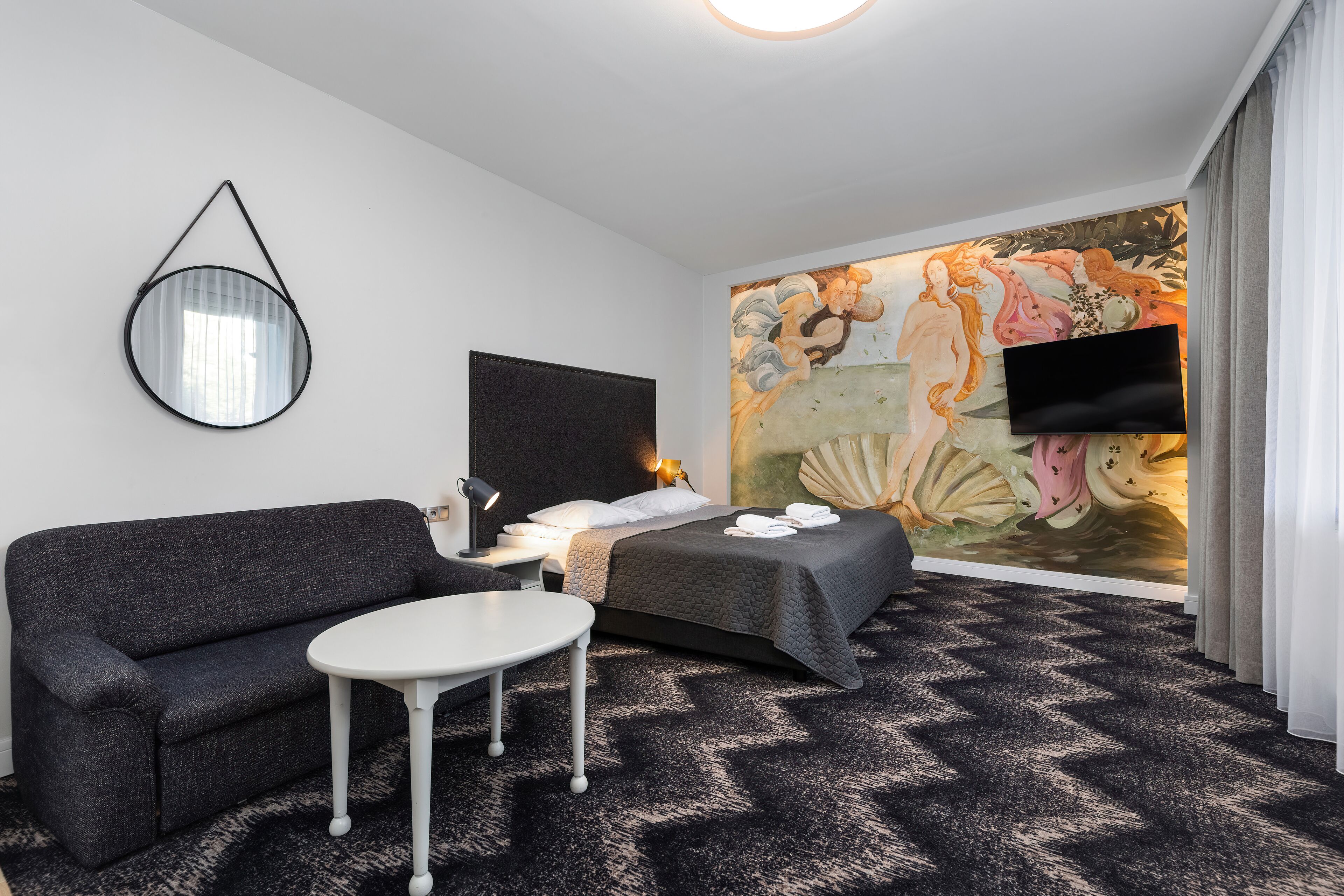 A hotel room with a bed, a sofa, a small table, a mirror, and a wall-mounted TV.
