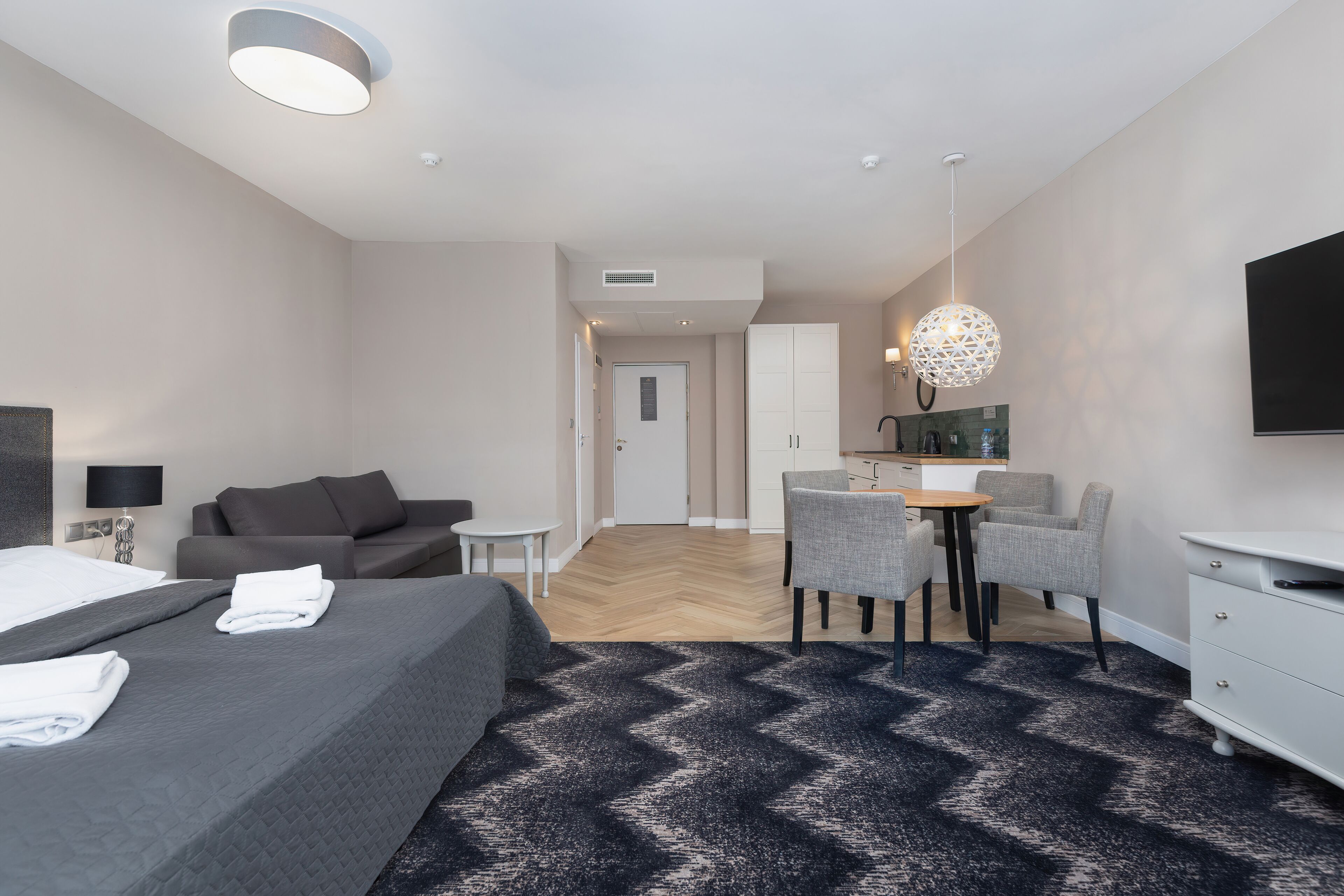 Ett modernt hotellrum med en säng, en soffa, en matplats med stolar, en TV och en mönstrad matta.