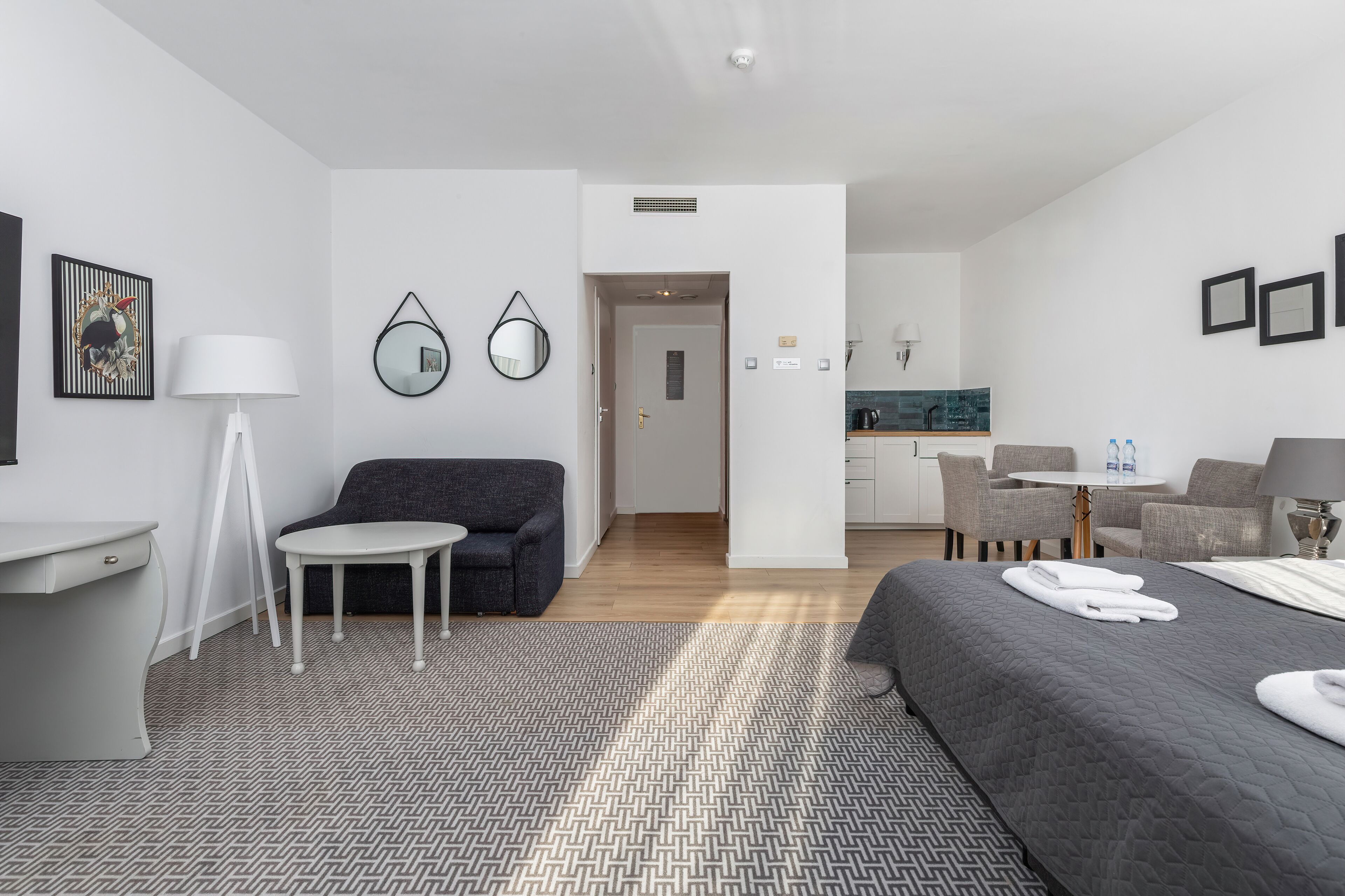 A modern hotel room with a bed, a desk, a chair, a small table, and a kitchenette in the background.