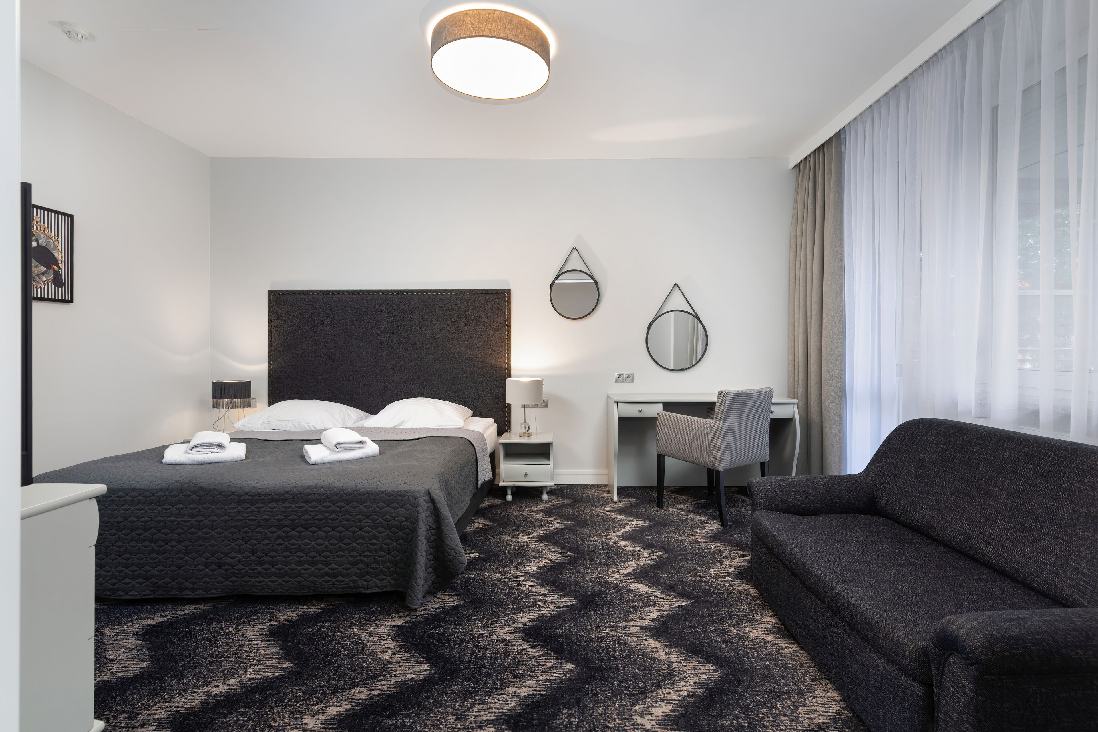 A hotel room with a bed, a desk, a chair, a sofa, and a large window with curtains.
