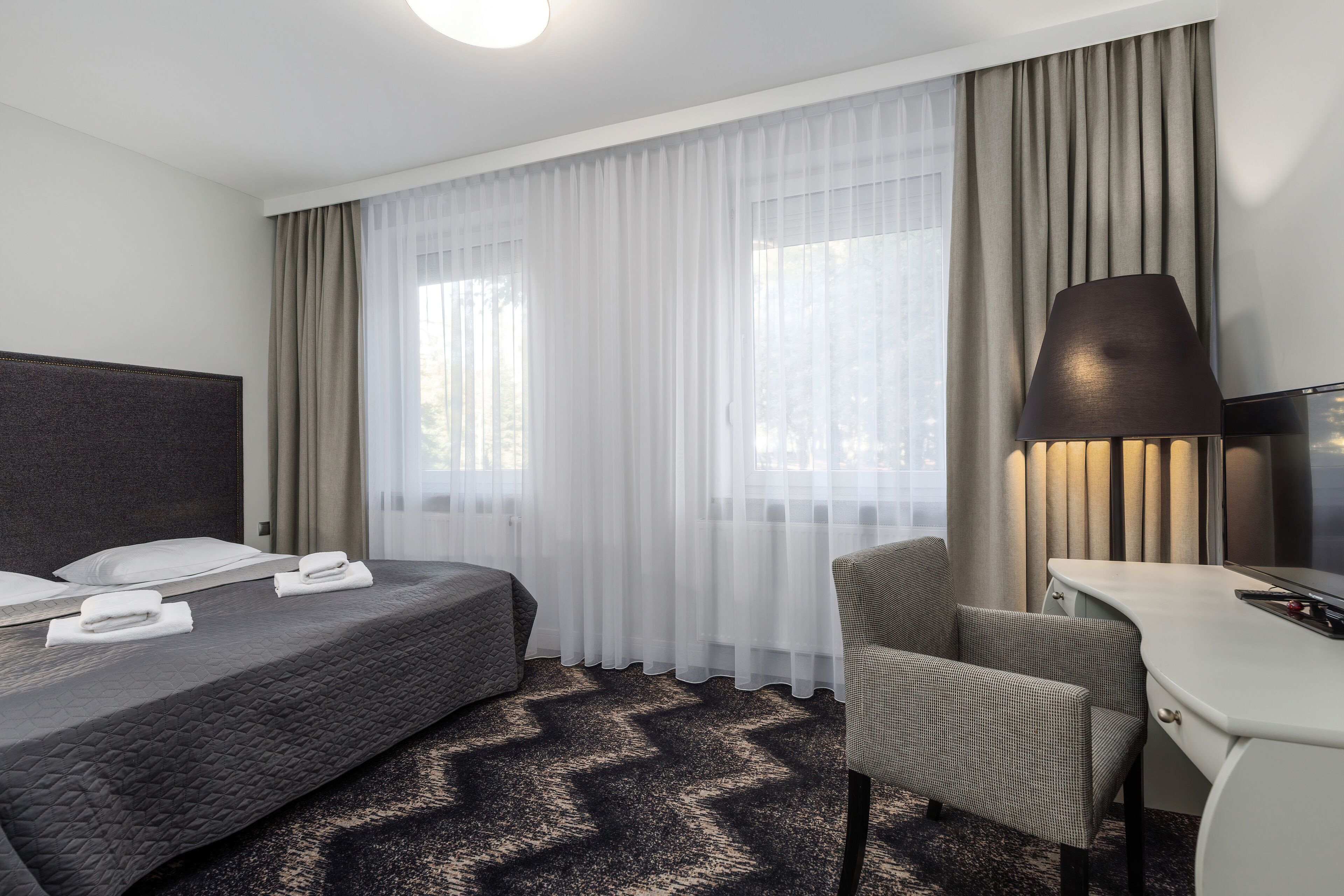 A hotel room with a bed, a desk, a chair, and a lamp.