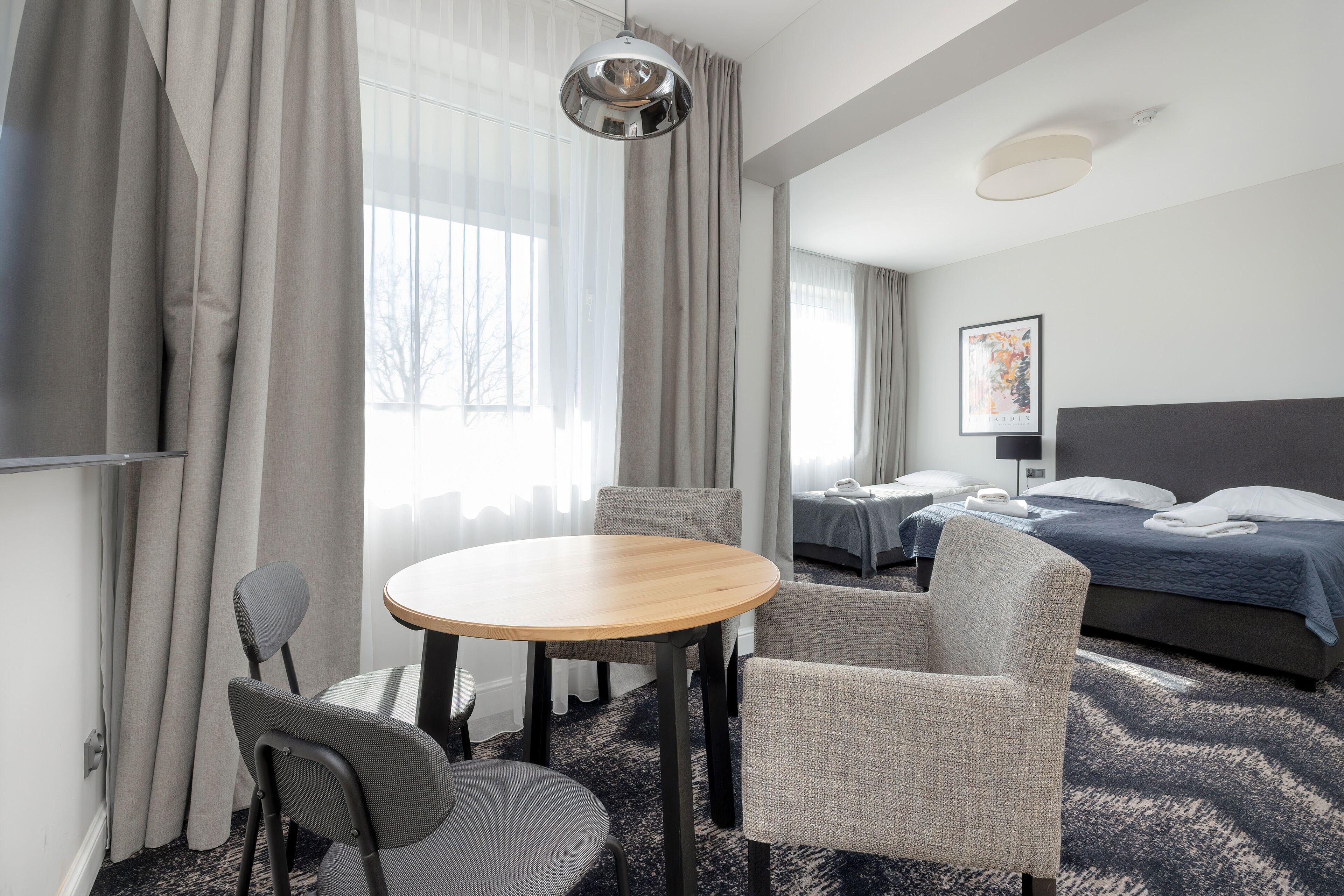 Een hotelkamer met een ronde houten tafel, twee grijze fauteuils, een bed met een blauwe dekbedovertrek en een ingelijst schilderij aan de muur.