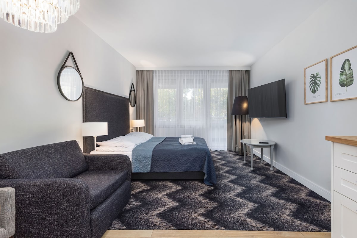 Een hotelkamer met een bed, een bank, een televisie, een lamp en wanddecoraties van planten in lijsten.