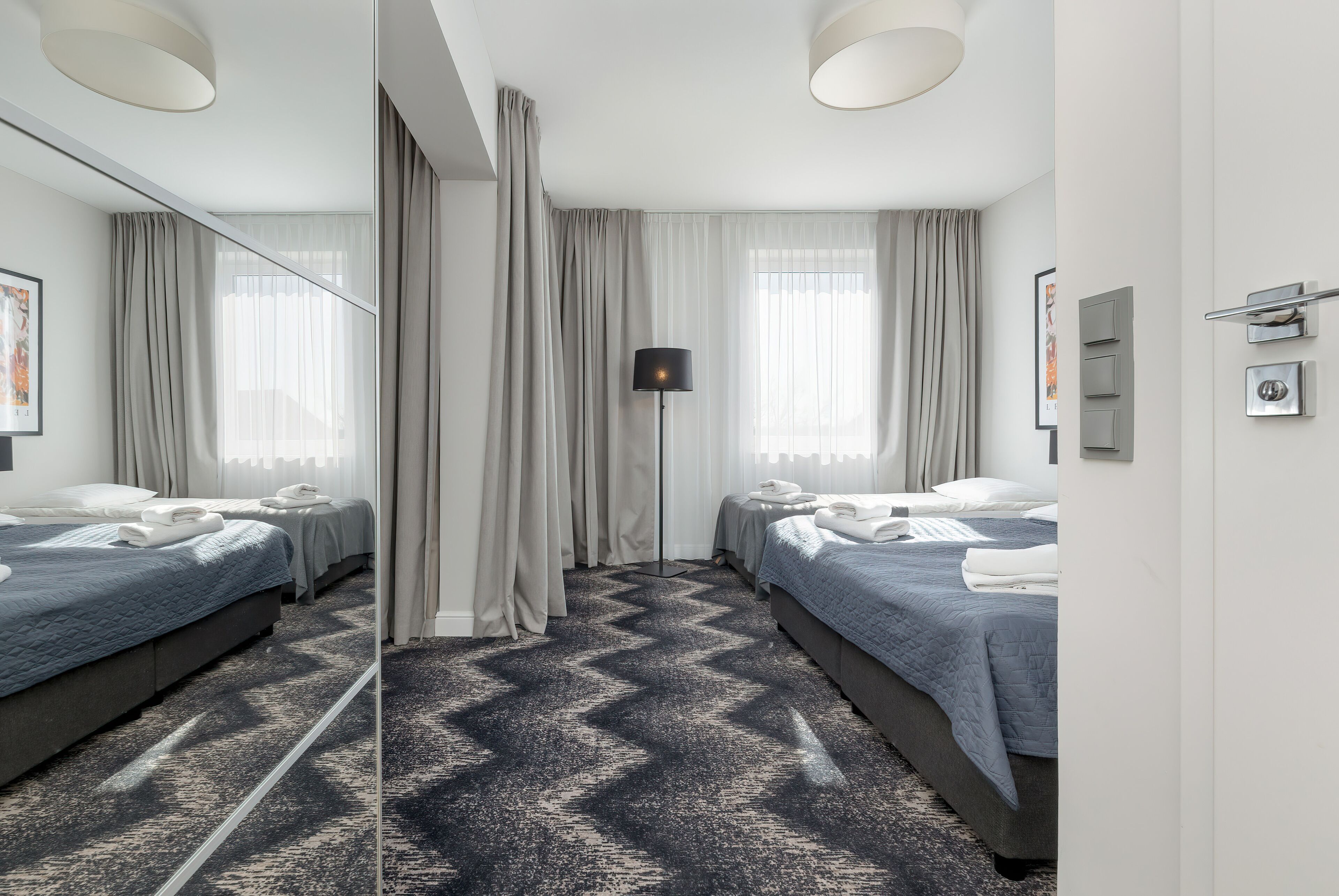 Habitación de hotel con dos camas, un espejo grande, una lámpara de pie y una alfombra estampada.