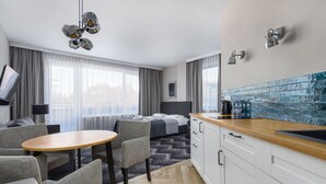 Une chambre d’hôtel moderne dotée d’un canapé, d’une table à manger, d’un coin cuisine et d’une grande fenêtre avec des rideaux.