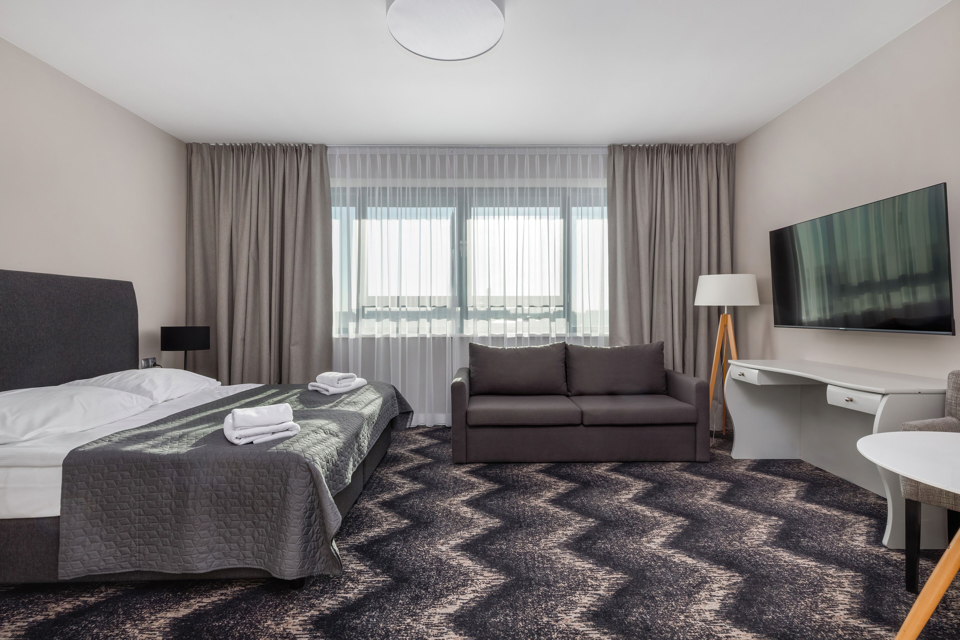 Hotelkamer met een bed, een slaapbank, een bureau, een televisie en een raam met gordijnen.