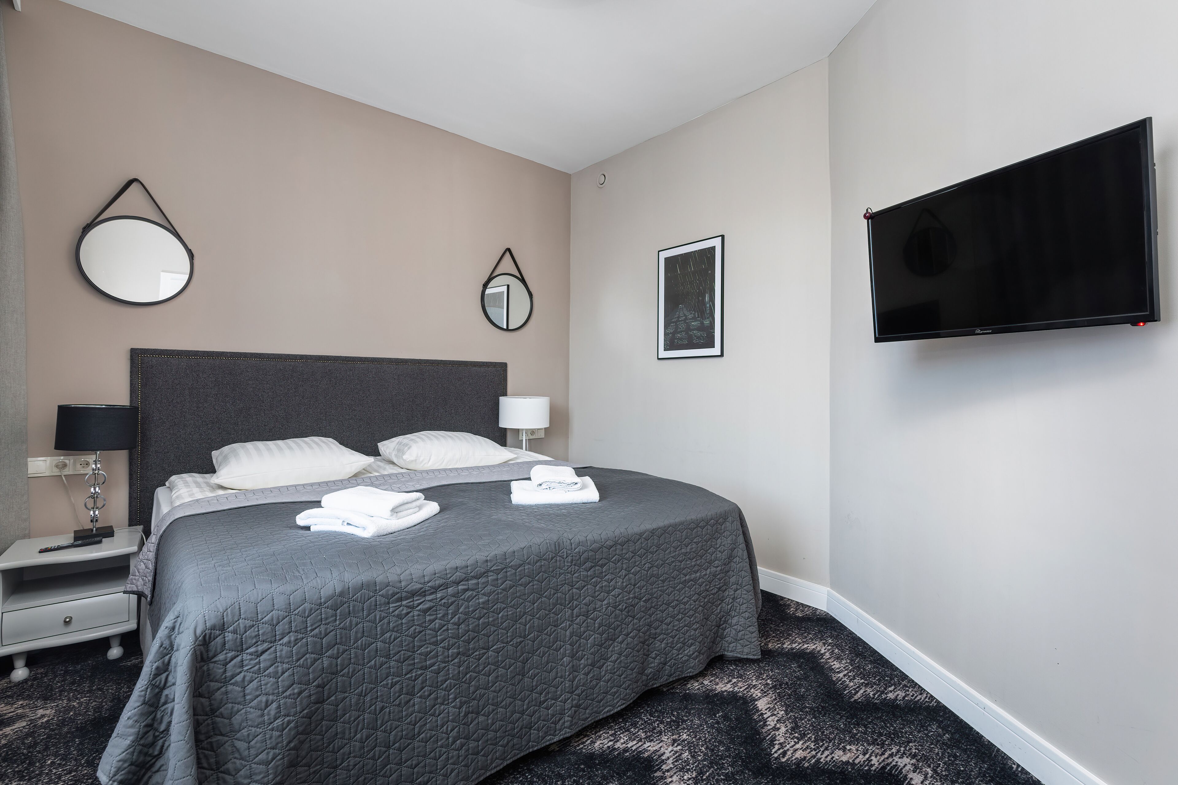 Ein ordentlich bezogenes Bett mit grauer Decke, ein Nachttisch mit Lampe und ein an der Wand befestigter Flachbildfernseher.