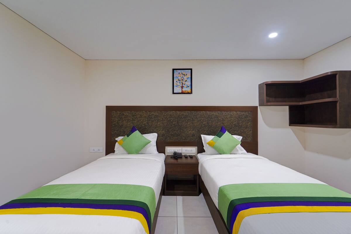 Habitación de hotel con dos camas individuales, una mesita de noche entre ellas, una pequeña mesa, un estante en la pared y un cuadro enmarcado.
