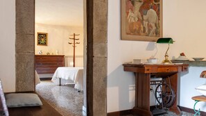A room with a wooden desk, a green desk lamp, a radiator, a bed with a white cover, a wooden dresser, and a painting on the wall.