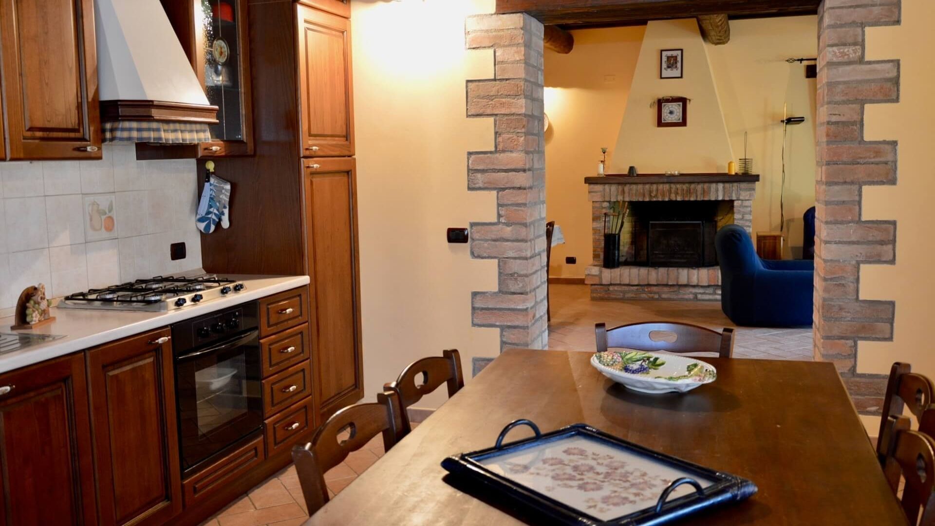A kitchen with wooden cabinets, a stove, and a dining table with chairs. There is a fireplace in the background.