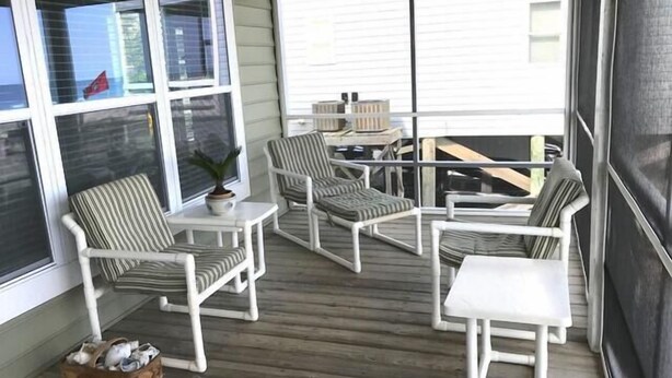 A covered outdoor seating area with white furniture and striped cushions, large windows, and a view of the surrounding area. - Holland House - Oceanfront, Dog Friendly (Pawleys Island)