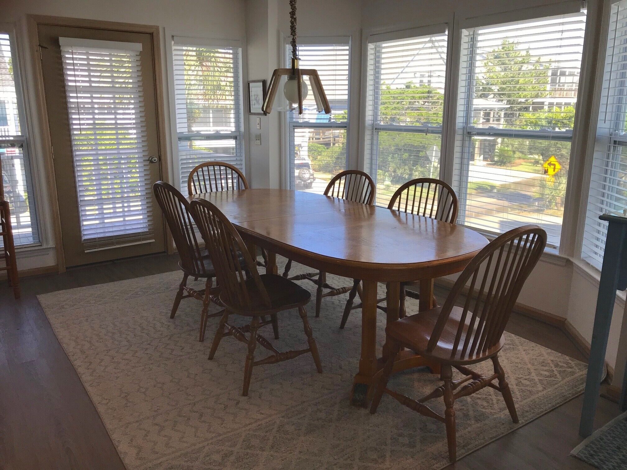 A dining room with a wooden table and chairs, large windows with blinds, and a hanging light fixture.