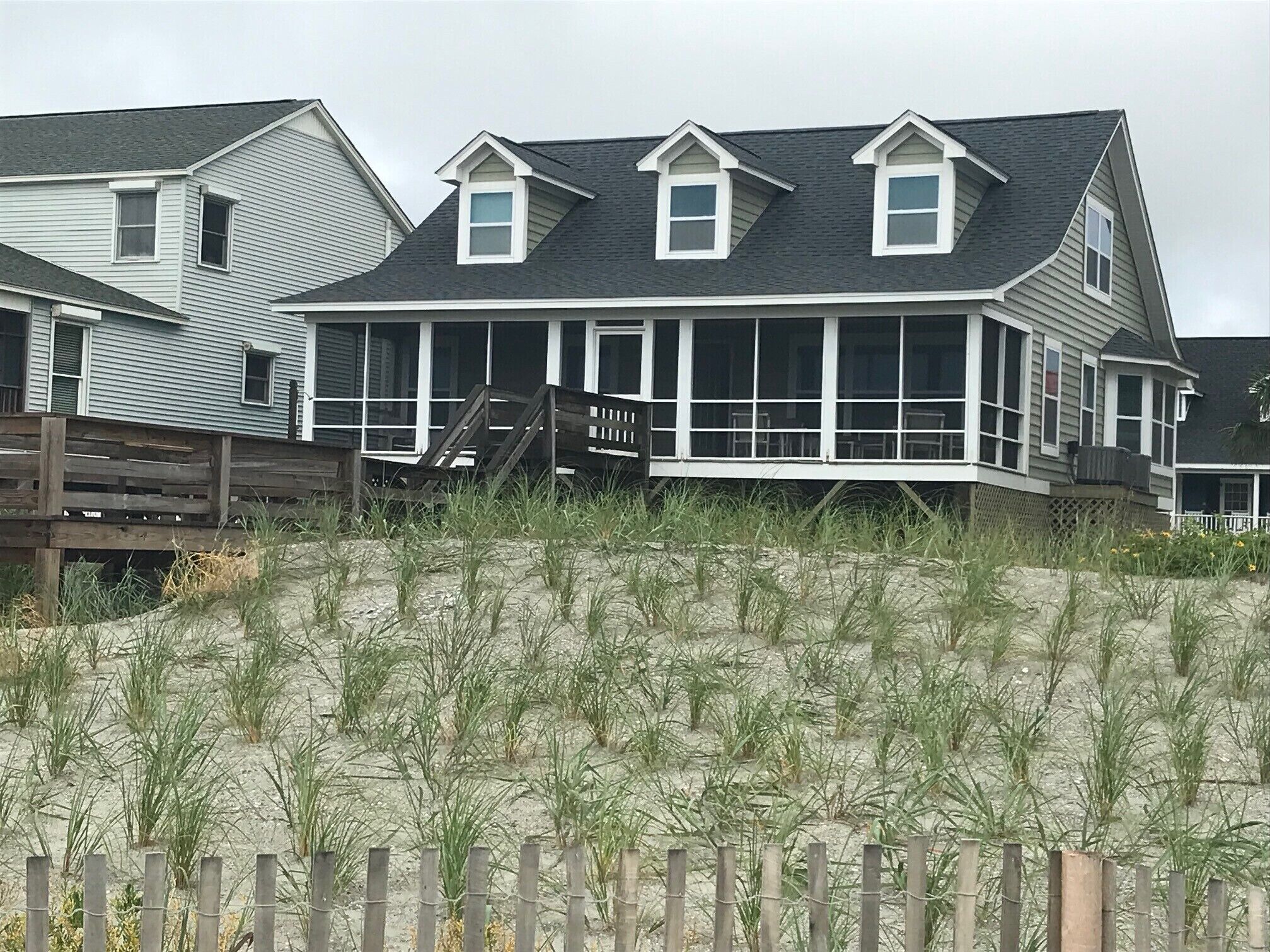 A beachfront house with a covered porch, a wooden deck, and a sandy beach in the foreground.