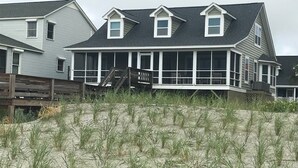 Et hus ved stranden med en overdækket veranda, en træterrasse og en sandstrand i forgrunden.
