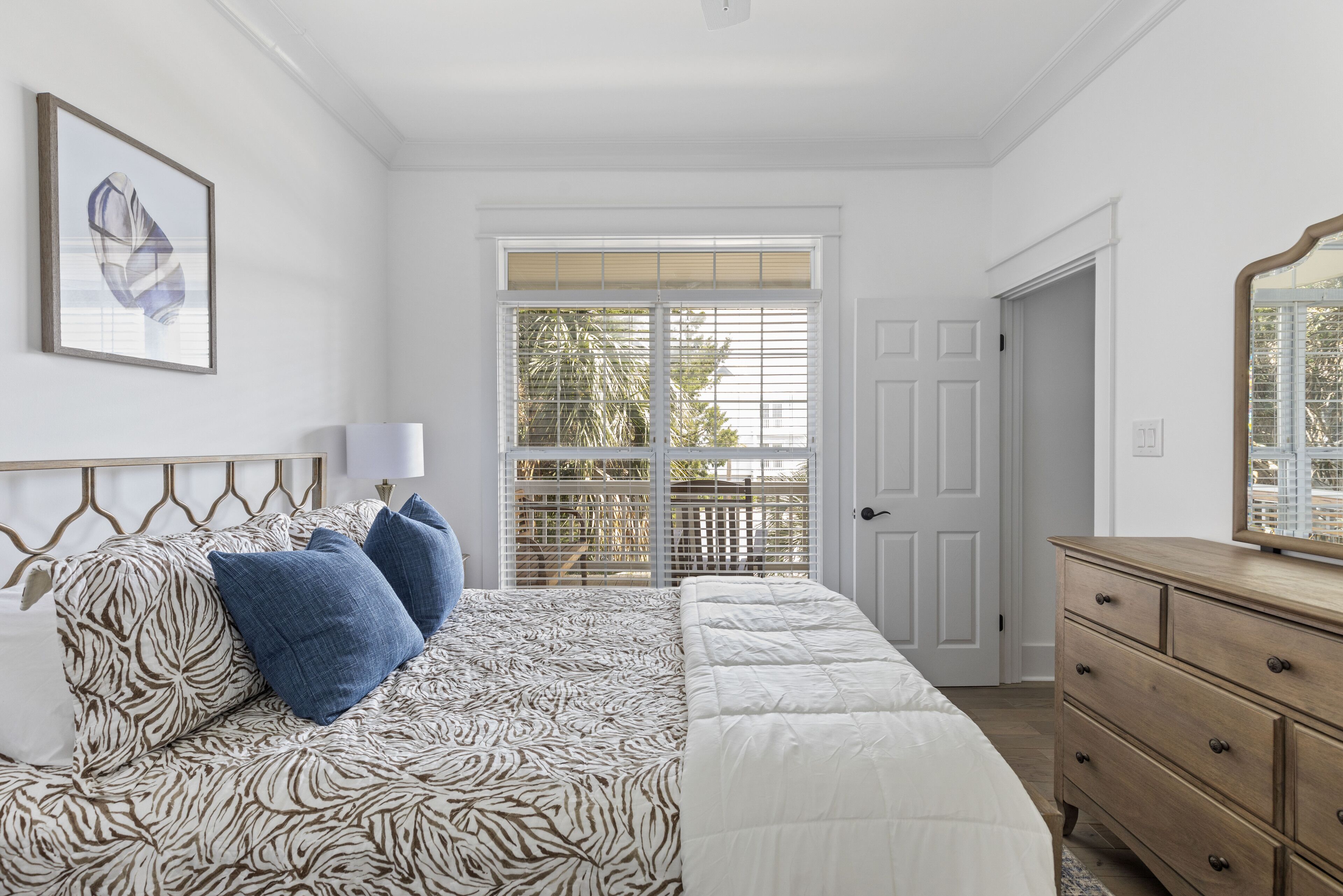 A bedroom with a bed, a dresser, a nightstand, a window with blinds, and a door.
