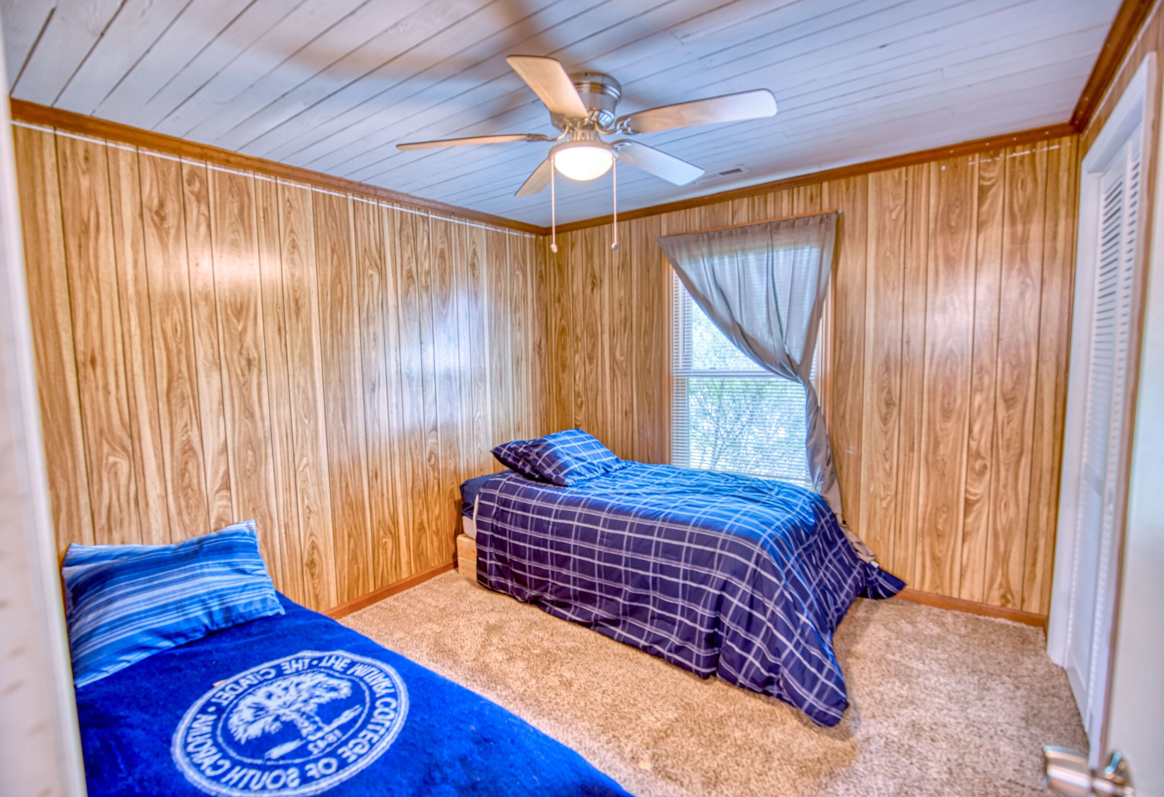 Ein Schlafzimmer mit Holzwandverkleidung, einem Deckenventilator, zwei Betten mit blauer Bettwäsche und einem Fenster mit Vorhängen.