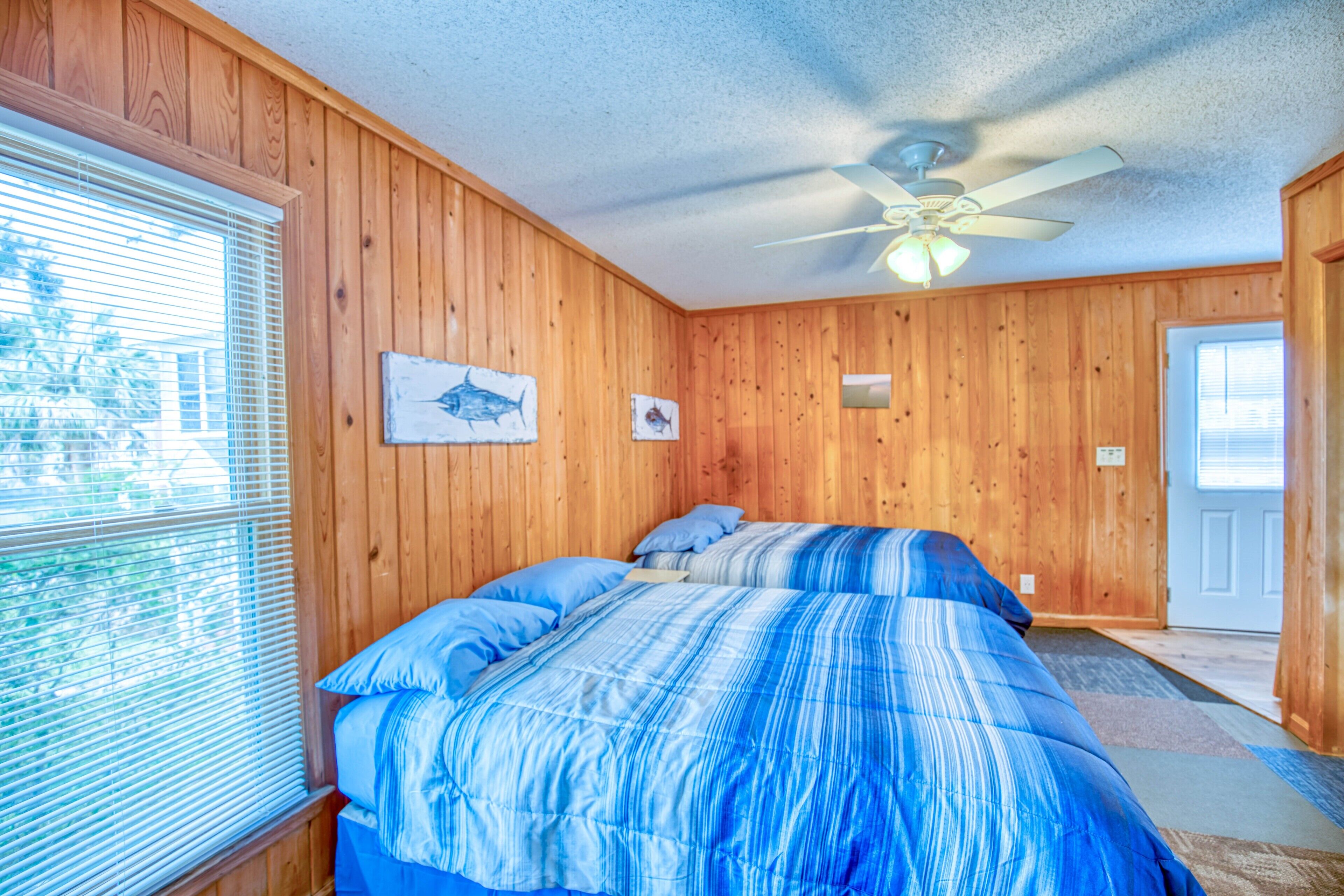 A bedroom with wooden walls, a bed with blue bedding, a ceiling fan, and a window with blinds.
