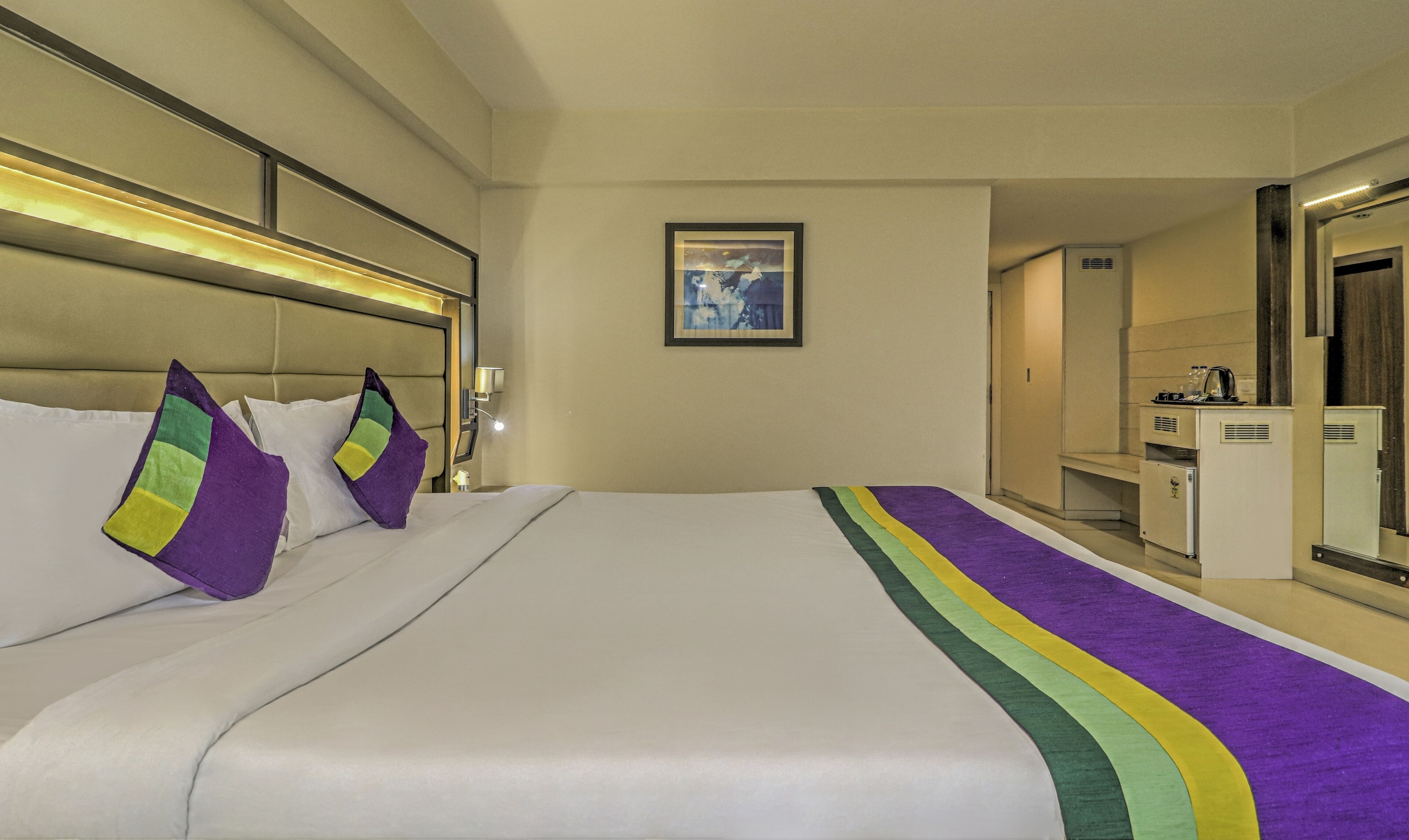 Habitación de hotel con una cama grande, una mesita de noche, un cuadro en la pared y una pequeña cocina.